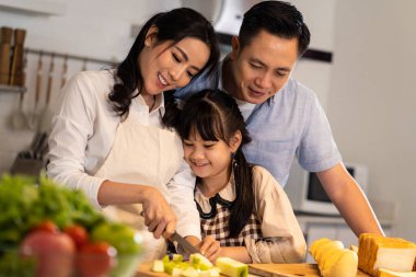 Asyalı mutlu aile evdeki mutfak odasında yemek hazırlıyor. Baba, anne ve çocuklar birlikte eğleniyorlar. Baba ve küçük kız yemek pişiren anneye bakıyor. Aile ilişkisi