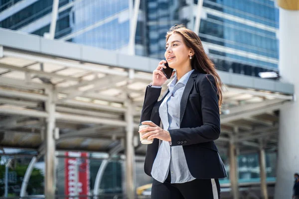 Asyalı genç ve zeki iş kadını şehirde telefonla konuşuyor. Elinde kahve fincanı olan kız gülen yüzle işe gidiyor. İş konsepti için telekomünikasyon ve mobil ağ.