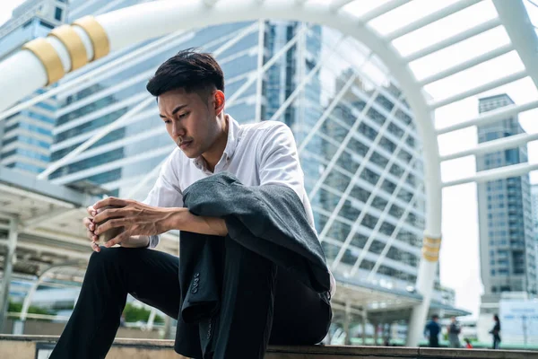 Asian business man feeling sad after unemployment from his company. The man sitting outdoor feeling stress and worry about losing the job due to world economic problem from covid-19 crisis concept.