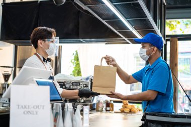 Covid-19 salgınından dolayı koruyucu maske takan Asya Yemekleri kuryesi kahve dükkanındaki restoran çalışanından içecek siparişi alıyor. Garson postacıya paket verir. Uzaktan alışveriş kavramı.