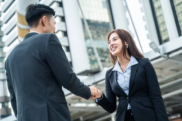 Asian businesswoman and man make handshake in the city building in the background. A partnership agreement is successful after complete the negotiation. Business deal, merger and acquisition concepts.