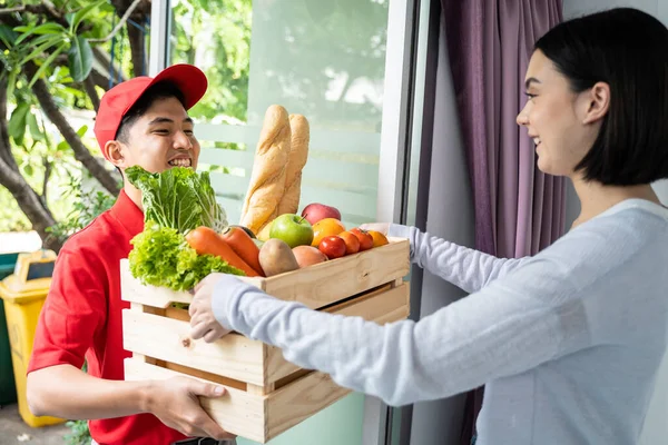 Active food delivery man working in red uniform deliver box of fruit and bread to young girl customer. Beautiful lady with smile face receiving groceries box from messenger infront of the door at home