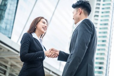 Asyalı işkadını ve erkek arka plandaki belediye binasında el sıkışırlar. Pazarlık tamamlandıktan sonra bir ortaklık anlaşması başarılı olur. İş anlaşması, birleşme ve satın alma kavramları.