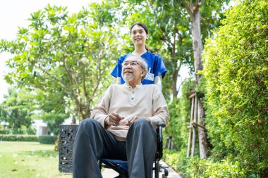 Asyalı mutlu emekli büyükbaba yeşil parkta genç kadın hemşireyle eğleniyor. Bakıcı doktor tekerlekli sandalyedeki yaşlı hastayı gülümseyerek itiyor, rahatlayın, şömine bakımının keyfini çıkarın..