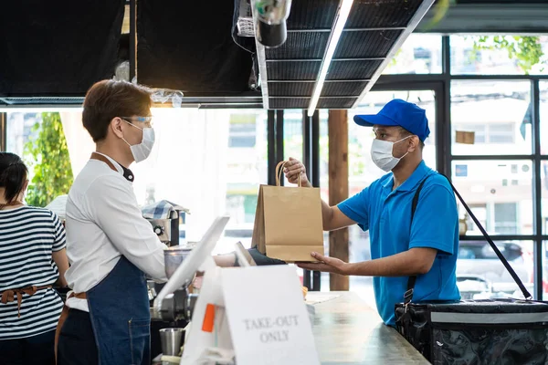 Asian Food deliverly man wearing protective mask due to Covid-19 pandemic, receive beverage order from restaurant worker at coffee shop. Waiter give takeaway bag to postman. Distance shopping concept.