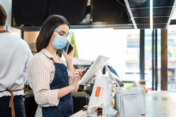 Asian cafe business owner wear protective face shield and mask due to Covid 19, use phone talk to customer and check order from touch screen on counter bar. Waiter prepare food for pick up, takeaway.