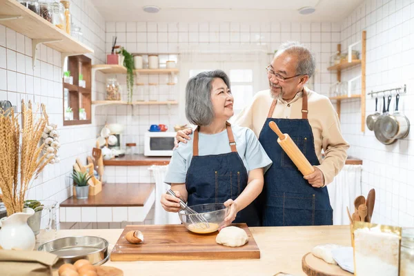 Portrait of Asian happy Senior elderly couple standing in kitchen at house feel happy and enjoy family retirement life together. Loving Older grandparent with apron, smiling and looking at each other.