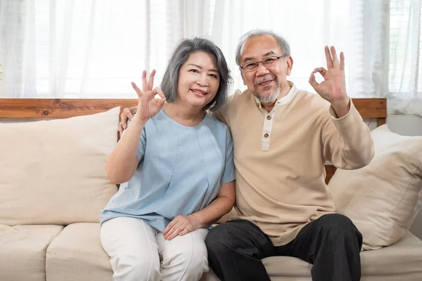 Asian happy family, Senior elderly couple enjoy retirement life at house. Older Grandfather and grandmother sit on sofa and smile, look at camera show okay gesture at home. Healthcare activity concept