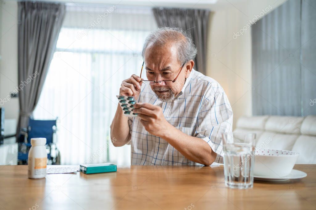 Asian Senior elderly male patient on eating table at nursing home care. Mature older man wear eyeglasses and look to medicine pill bottle and read prescription and content. Medical insurance concept.