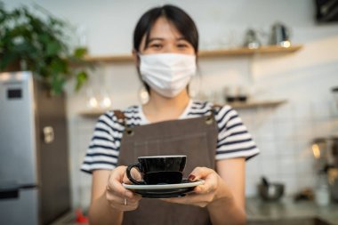 Portre Asyalı Garson Covid19 salgınından dolayı maske takıyor, kafede iki eli olan müşteriye bir fincan sıcak kahve veriyor. Önlüklü bir işyeri sahibi restoranda çalışıyor. Gülümse ve kameraya bak..