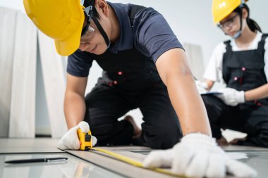 Asyalı zanaatkar veya marangoz erkek ve kadın güvenlik kaskı ve gözlük takıyor, koridorda parke döşeyerek çalışıyorlar. Katılımcı İnşaatçı, apartman veya evin yenilenmesi için zemine kaplama döşeme kurar.
