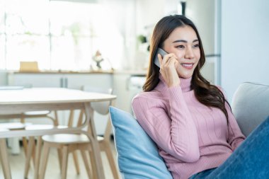 Asyalı güzel kadın kanepede oturur ve cep telefonuyla konuşur. Mutlu çekici genç kız evde vakit geçirir, rahatlar, iletişim kurar ve oturma odasında akıllı telefondan tartışır..