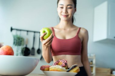 Asyalı çekici aktif kadın mutfaktan yeşil bir elma alıyor. Spor giyim sektöründeki güzel spor kızı meyve seçme ve tatlı çöreği görmezden gelme arzusunda. Egzersiz yaptıktan sonra sağlıklı beslenmenin tadını çıkar.