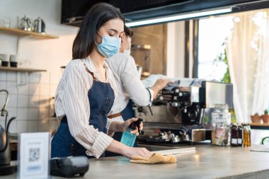 Kafe garsonu maske takıyor, mağazayı açmak için dezenfektanlı temiz bir restoran. Asya kökenli genç çift, Covid19 kilitlenmesinden sonra dükkanı yeniden açmadan önce restoranda dezenfekte, temizleme ve ovalama yapıyor..