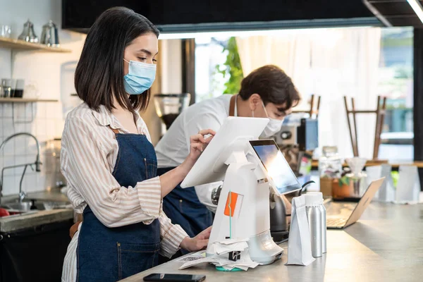 Asian cafe business wear mask, use laptop check food order and payment. Young attractive beautiful waiter use touch screen on counter bar to prepare food for pick up, takeaway during Covid 19 pandemic