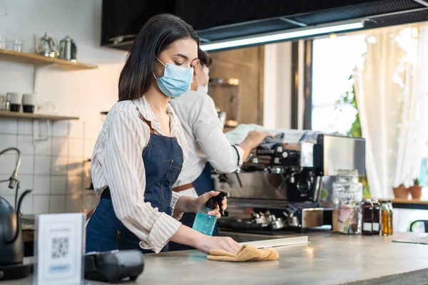 Cafe waiter wear mask, clean restaurant with sanitizer to open store. Asian attractive young couple disinfecting, wiping and rubbing on counter in restaurant before reopen store after Covid19 lockdown
