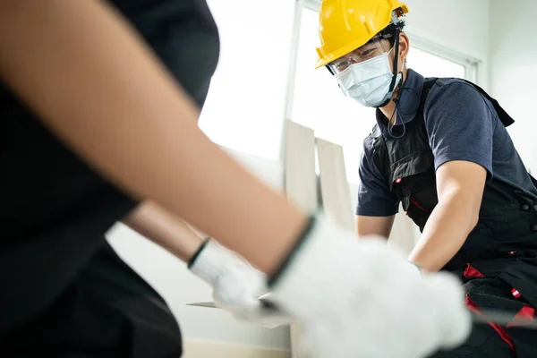 Asian builder worker people wear mask installs laminate board on floor. Construction team or Carpenter man and woman wear safety helmet work to repair parquet on corridor to renovate home and housing.