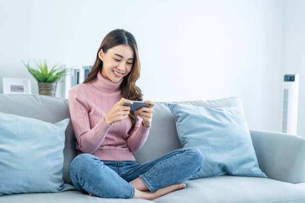 Азиатская молодая красивая женщина играет в мобильную игру на смартфоне дома. Привлекательная случайная девушка чувствовать себя счастливой и расслабиться, сидеть на диване весело сенсорный экран на телефоне, чтобы играть видео наслаждаться победой в доме