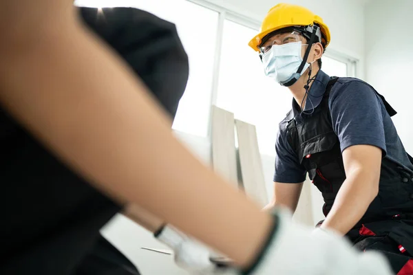 Construction worker installs laminate board on floor to renovate house. Asian builder man and woman wear safety helmet and protective face mask due to Covid-19 pandemic while work to repair apartment.