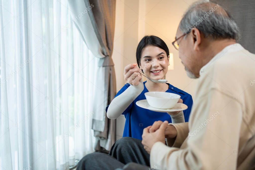 Asian young woman nurse at nursing home taking care of disabled senior elderly man on wheelchair. Beautiful Therapist doctor feeding food to older patient in house. Medical insurance service concept.