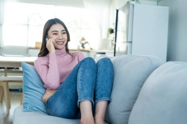 Asyalı güzel kadın kanepede oturur ve cep telefonuyla konuşur. Mutlu çekici genç kız evde vakit geçirir, rahatlar, iletişim kurar ve oturma odasında akıllı telefondan tartışır..