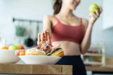 Asyalı çekici aktif kadın mutfaktan yeşil bir elma alıyor. Spor giyim sektöründeki güzel spor kızı meyve seçme ve tatlı çöreği görmezden gelme arzusunda. Egzersiz yaptıktan sonra sağlıklı beslenmenin tadını çıkar.