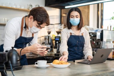 Asyalı barista çifti kafe restoranında dijital pazarlama yapmayı sever. İş ortakları mutlu hisseder, kahve fotoğrafı çeker ve sosyal ağ için dizüstü bilgisayarı kullanır.