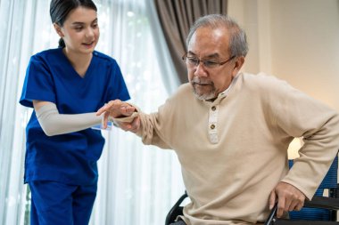 Asyalı bakıcı kız doktor yaşlı engelli adamın eve girişini destekliyor. Yaşlı ve olgun bir hastayla ilgilenen çekici bir hemşire. Evde tekerlekli sandalyede oturuyor ve terapi görüyor..