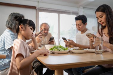 Asyalı Görmezden Gelen Aile yemek yerken küçük çocuğa dikkat etmiyor. Üzgün kız yemek masasında oturmuş akıllı telefon kullanan bağımlı anne ve babalara bakıyor. Evde kötü ebeveynlik.