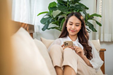 Asyalı güzel kadın kanepede oturur ve cep telefonuyla sohbet eder. Mutlu çekici genç kız evde vakit geçirir, rahatlar, iletişim kurar ve oturma odasında akıllı telefondan tartışır..