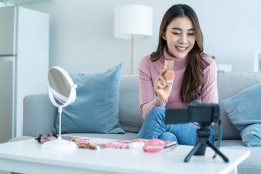 Evdeki kozmetiği incelemek için kamerayla Asyalı kadın video blogcu konuşması. Güzel Güzellik blogcusu Nüfuzlu Kız kanepeye otur ve canlı yayın yap, vakıf şovu yap, evdeki takipçilere makyaj yap.