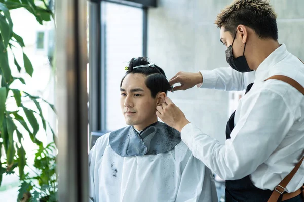 Asian hairdresser barber wear mask, cut hair of attractive man in shop. Handsome young man customer in modern salon barbershop, enjoy getting haircut service by stylist man during Covid 19 pandemic.