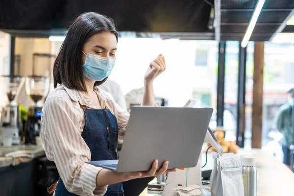 Asian cafe shop owner woman check sale order from laptop on counter. Young girl restaurant waiter wear mask due to Covid19, look at computer and happy for income. Entrepreneur and Economic of business