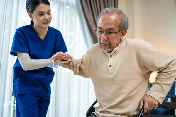 Asyalı bakıcı kız doktor yaşlı engelli adamın eve girişini destekliyor. Yaşlı ve olgun bir hastayla ilgilenen çekici bir hemşire. Evde tekerlekli sandalyede oturuyor ve terapi görüyor..