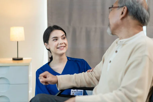 Asian nurse take care of senior man on wheelchair at nursing home care. Attractive happy Caregiver girl doctor greeting and talking with older elderly mature disabled patient male in hospital ward.