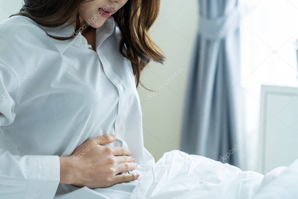 Asian beautiful sick girl in pajamas getting up from sleep in bedroom. Attractive young woman feel bad and painful after wake up on bed, having stomach ache or periods then clasping belly in morning.