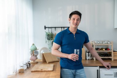 Asyalı çekici bir adam mutfakta bir bardak temiz su tutuyor. Genç, susamış yakışıklı erkek, sabahın erken saatlerinde sağlık hizmetleri için bardağa temiz mineral döküp içiyor.