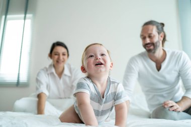 Gülümseyen çocuk arka planda ebeveynleriyle yatakta oynuyor.