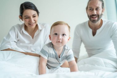 Gülümseyen ebeveynler beyaz yatakta emekleyen bebeği izliyor.