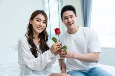 Asyalı genç adam sürpriz yaptı ve gül çiçeğini güzel kız arkadaşına verdi. Çekici, romantik, yeni evli çift yatakta oturan erkek ve kadın yıldönümünü ve sevgililer gününü birlikte kutluyorlar.