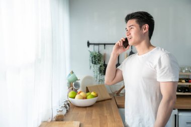 Genç Asyalı adam akıllı telefonla konuşuyor ve mutfakta dururken gülümsüyor..