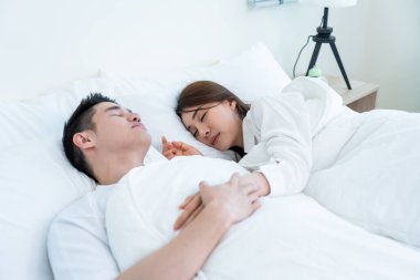 Asian attractive young couple lying down on bed to sleep in bedroom. Beautiful new marriage man and woman in pajamas feel happy and relax on comfortable pillow in the early morning together in house.