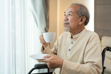 Tekerlekli sandalyede oturan yaşlı bir adam elinde fincan ve çay tabağı tutuyor.