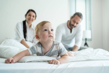 Anne babalar arka planda gülümserken bebek yatakta yatıyor.