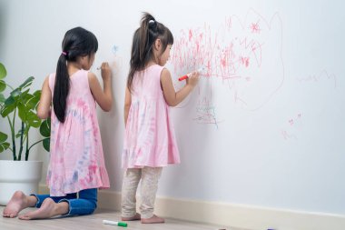 Asian young sibling kid girl enjoy paint on white wall in living room. Little adorable children having fun drawing and coloring art picture with hapiness enjoy creativity activity on holiday at home.