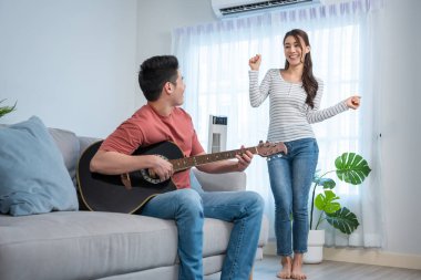 Kadın oturma odasında dans ederken, erkek gitar çalıyor.