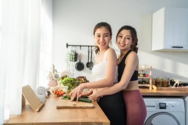Asyalı iki genç kadın, mutfakta spor kıyafetleri içinde salata pişiriyorlar..