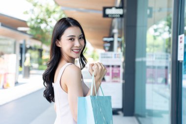 Elinde alışveriş poşetleri olan gülümseyen bir kadın dükkânın yakınında yürüyor.