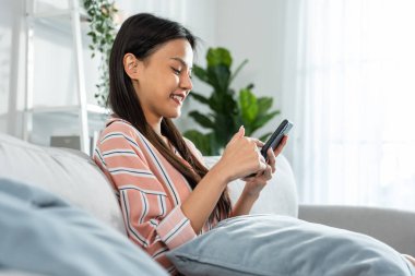 Asyalı güzel kadın kanepede oturur ve cep telefonuyla sohbet eder. Mutlu çekici genç kız evde vakit geçirir, rahatlar, iletişim kurar ve oturma odasında akıllı telefondan tartışır..