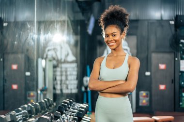 Spor giyimli Afrikalı spor kızın portresi spor salonunda egzersiz yapıyor. Çekici genç bayan sporcu spor salonu veya fitness kulübünde makine ve ekipmanlarla antreman yaptıktan sonra kollarını kavuşturup duruyor..